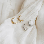 Two pairs of Eclipse Moon Studs, one in gold and one in silver, elegantly displayed on white fabric.
