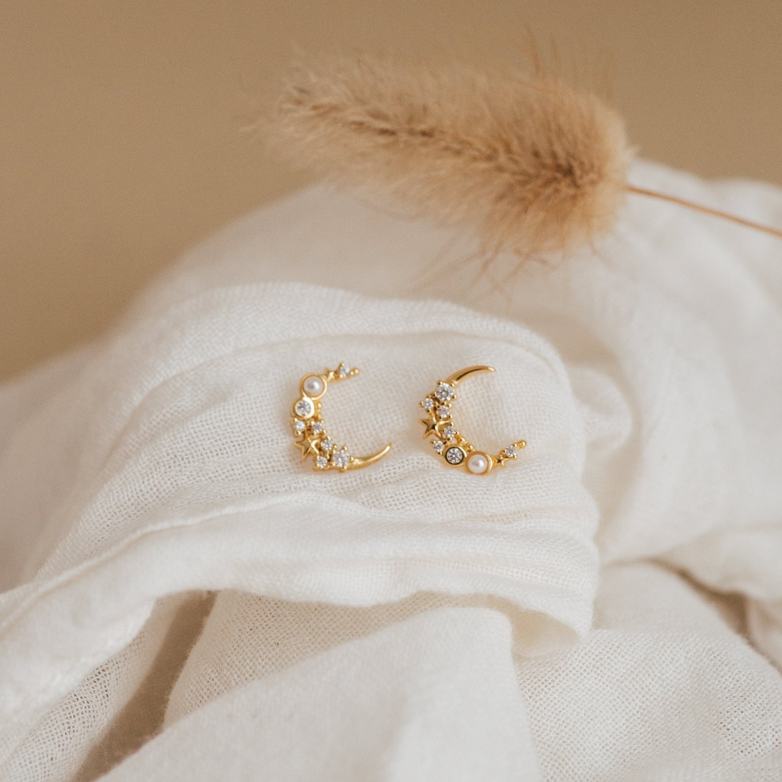Eclipse Moon Studs—gold crescent earrings with sparkling crystals—are displayed on soft white fabric, accented by dried grass in the background.