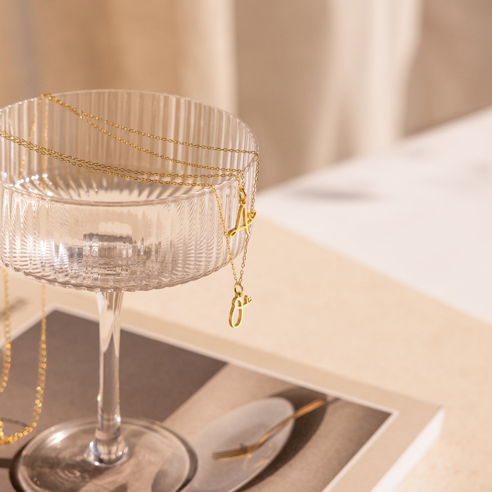 The Letter Heart Necklace, a delicate gold piece, drapes over a glass cup resting on a book whose cover features a plate and fork.