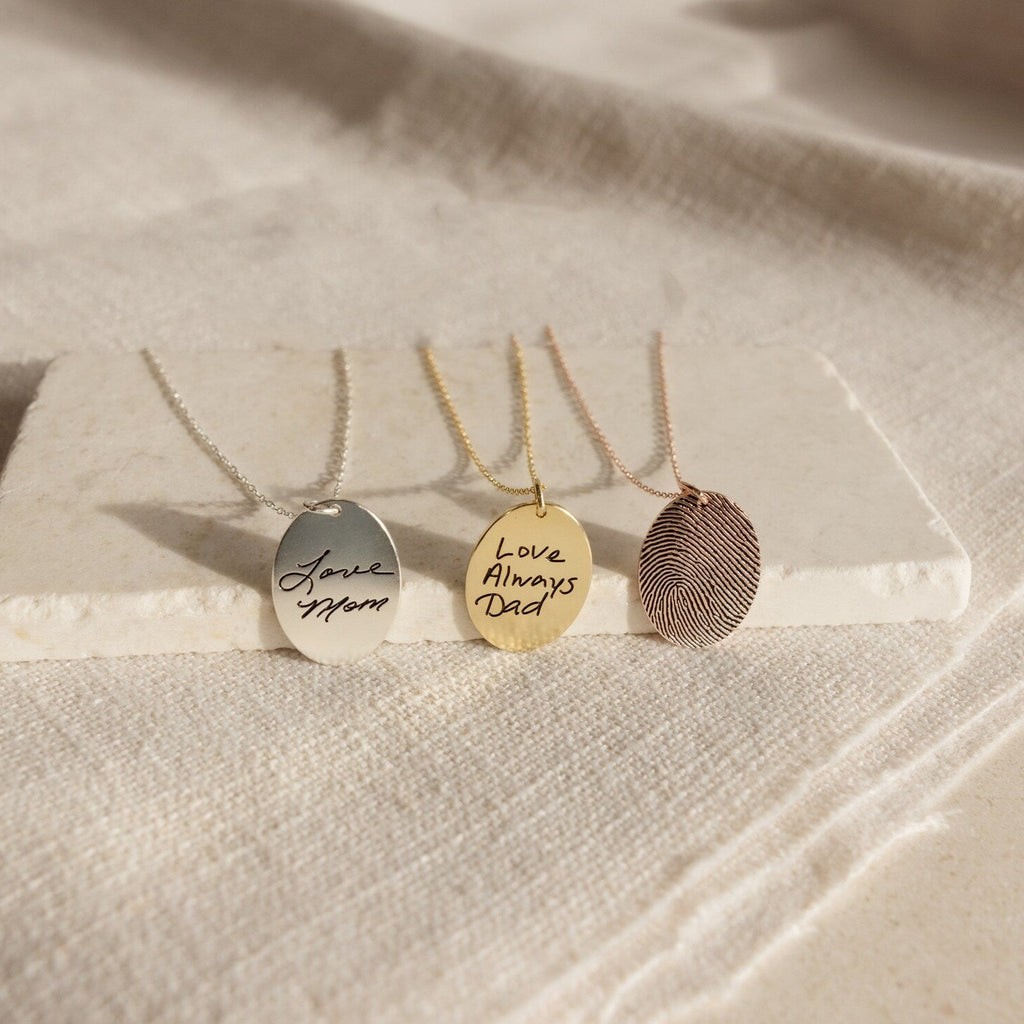 Three engraved necklaces rest on a stone slab: Love Mom, Love Always Dad, and the Oval Fingerprint Necklace—each crafted as meaningful memorial jewelry with personalized etching.
