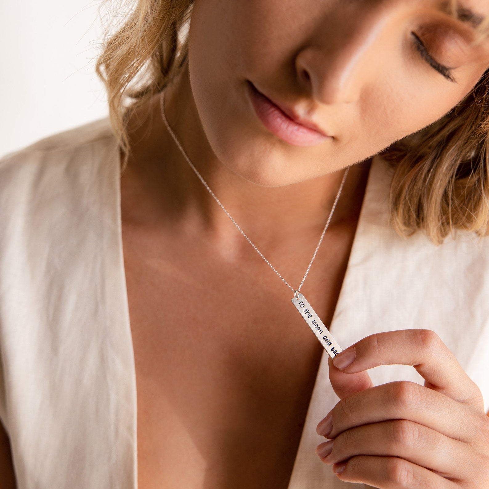 A woman wearing a cream top holds the Vertical Handwriting Bar Necklace with engraved text close to her chest.