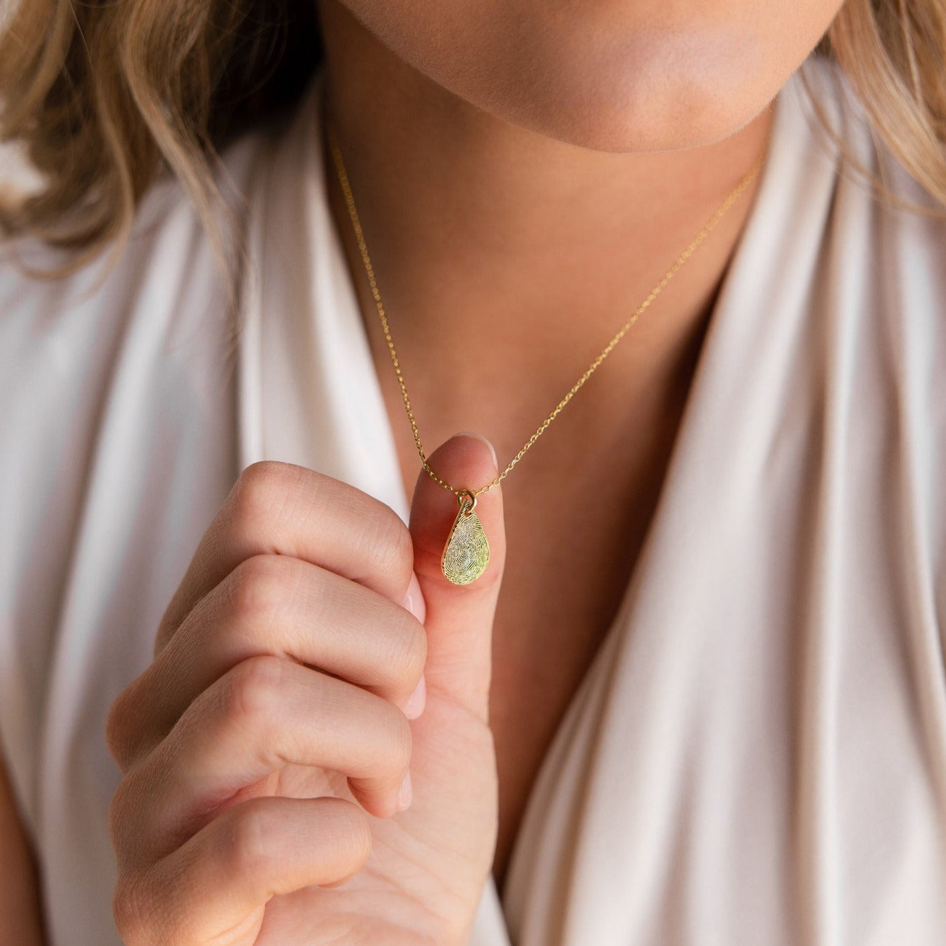 Woman wearing a light sleeveless top holds the Tear Drop Fingerprint Necklace, featuring a gold teardrop pendant adorned with a green stone.