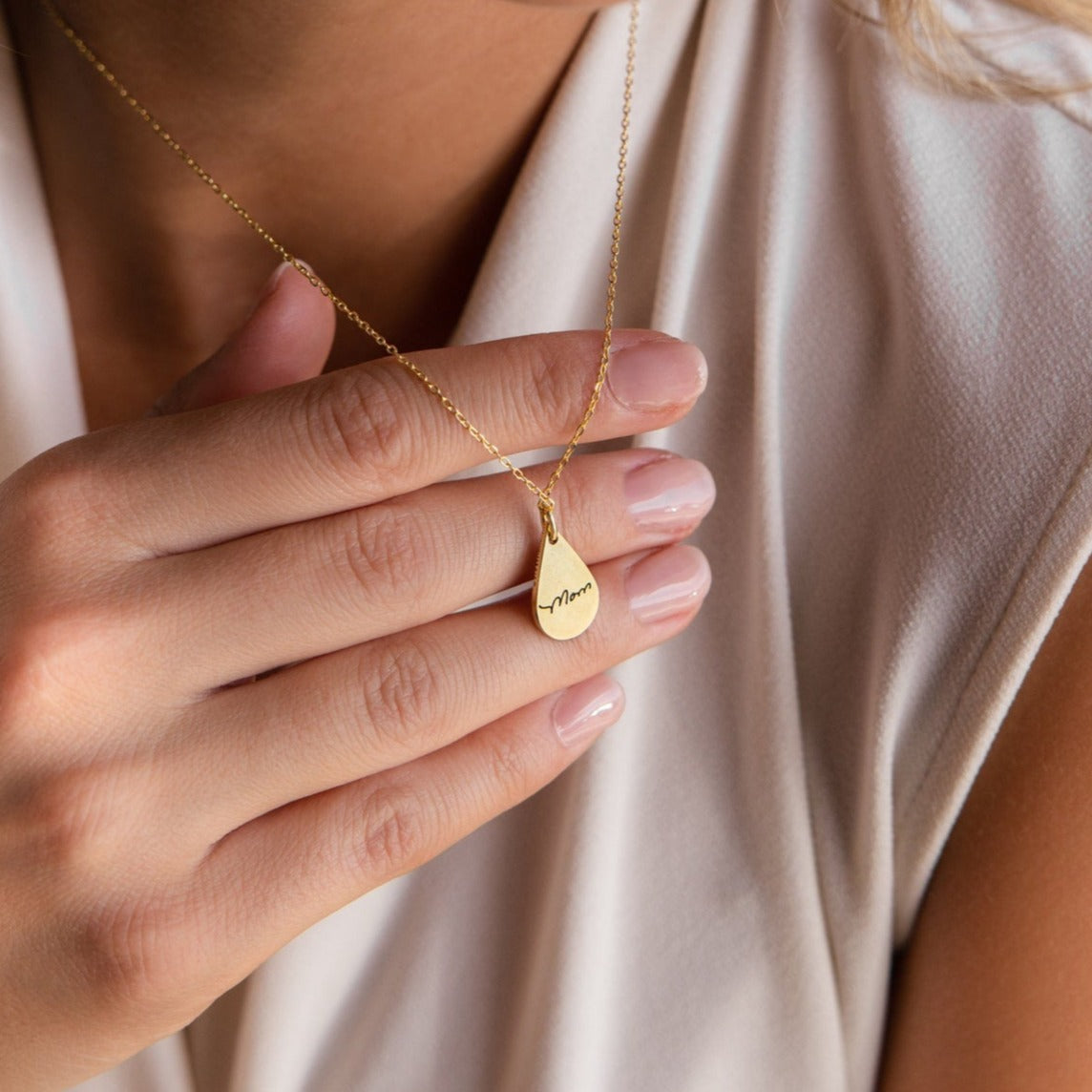 A hand holds the Tear Drop Fingerprint Necklace—a gold teardrop-shaped pendant engraved with "Mom"—against a cream blouse.