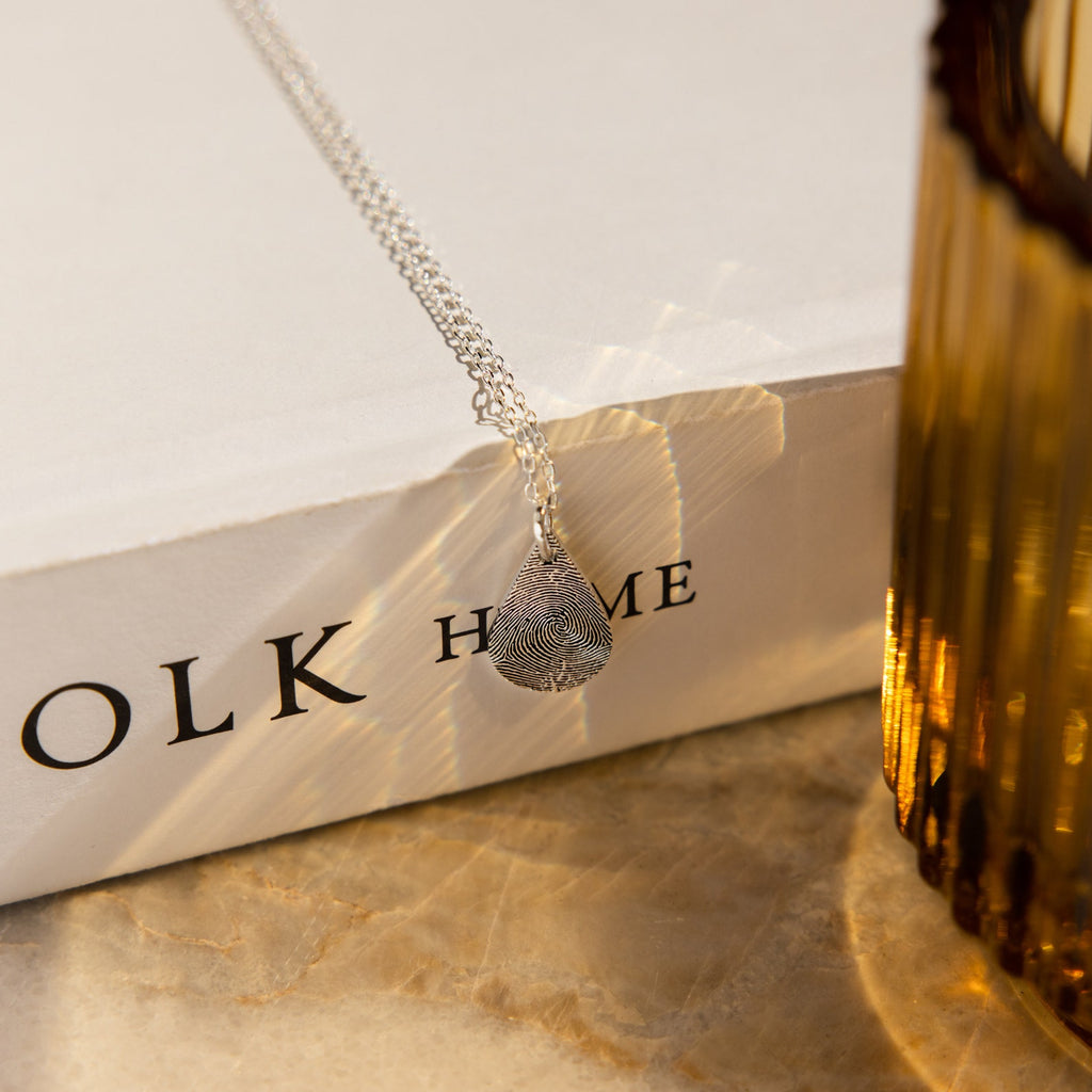 The Tear Drop Fingerprint Necklace, a silver teardrop-shaped pendant featuring a fingerprint keepsake, rests on a white book next to an amber glass.