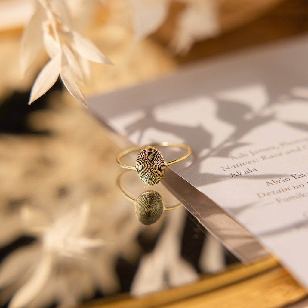 The Fingerprint Oval Ring, a handcrafted gold piece featuring a unique fingerprint design, is displayed on a reflective surface beside white paper.