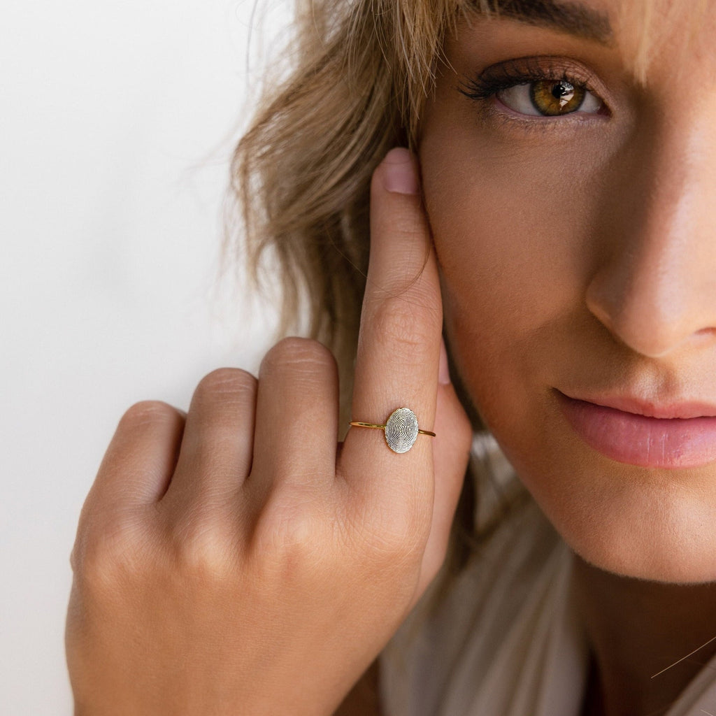A woman with light brown hair gently touches her face, showcasing the handcrafted Fingerprint Oval Ring featuring a unique white stone.