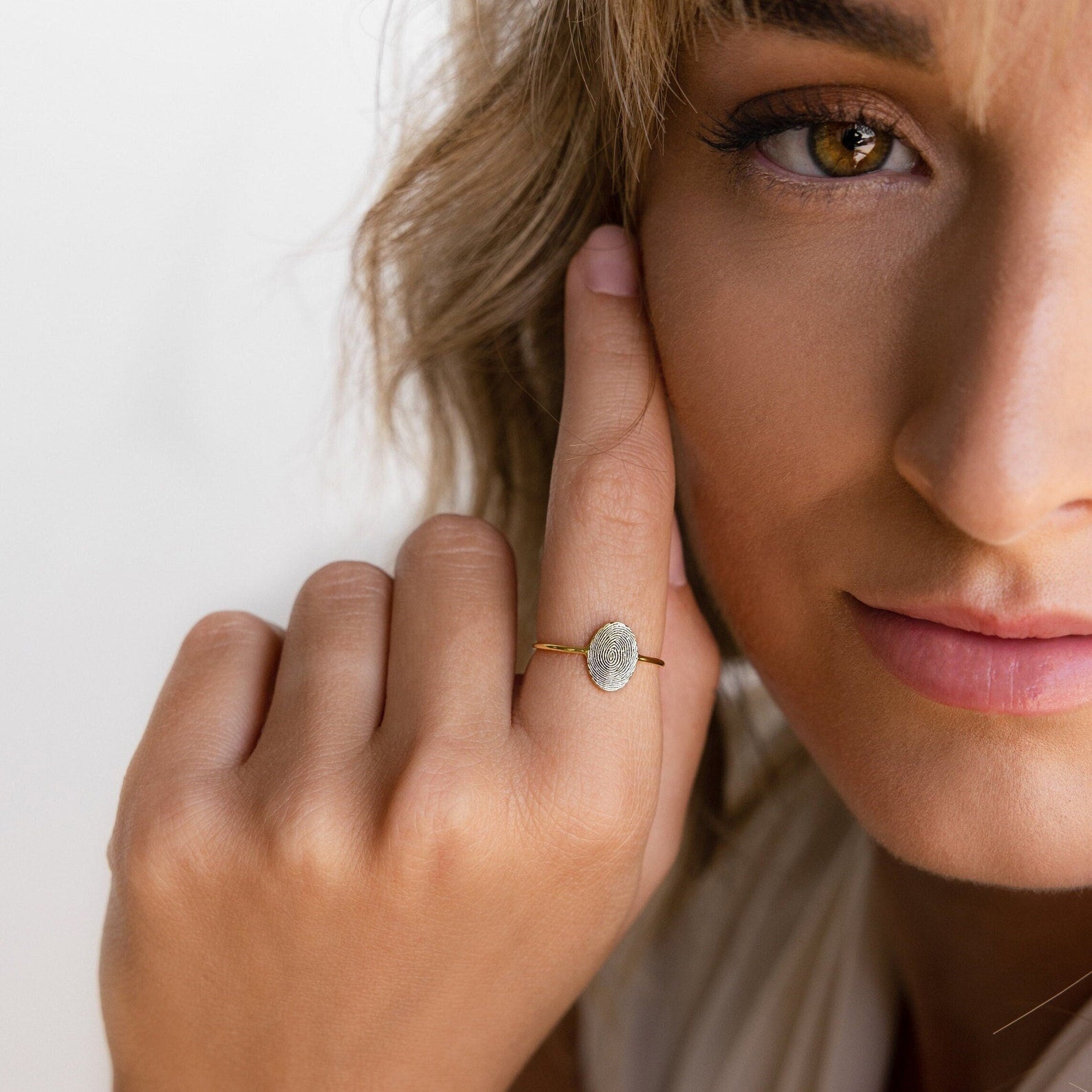A woman with light brown hair gently touches her face, showcasing the handcrafted Fingerprint Oval Ring featuring a unique white stone.
