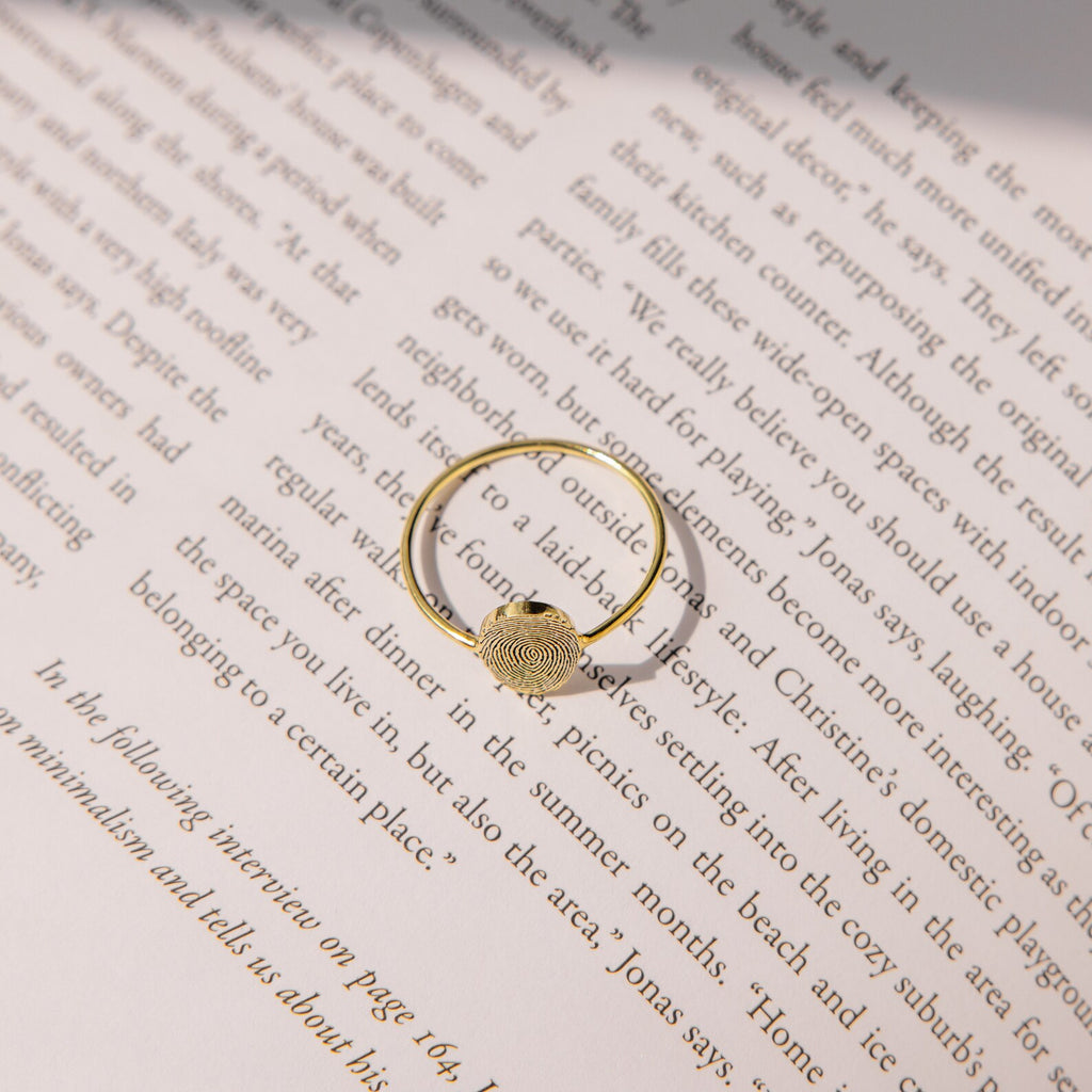 The Fingerprint Oval Ring, a handcrafted gold piece featuring a unique fingerprint design, is displayed on the open pages of a book under gentle lighting.