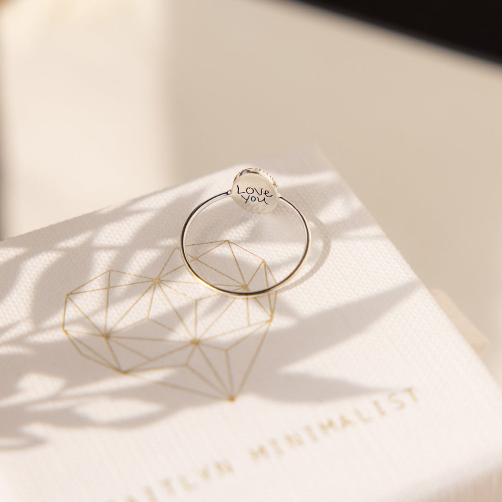 A handcrafted Fingerprint Oval Ring rests on a white box featuring a geometric heart and gold lettering.