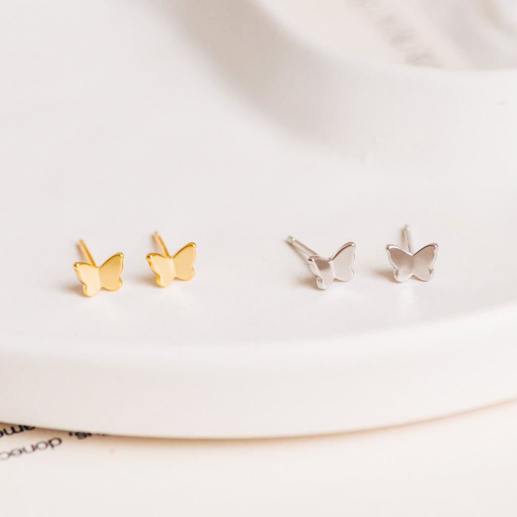 The Emilia Butterfly Studs feature two pairs of dainty butterfly earrings—one gold, one silver—displayed on a white surface, perfect for adding a trendy touch to any outfit.
