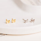 The Emilia Butterfly Studs feature two pairs of dainty butterfly earrings—one gold, one silver—displayed on a white surface, perfect for adding a trendy touch to any outfit.