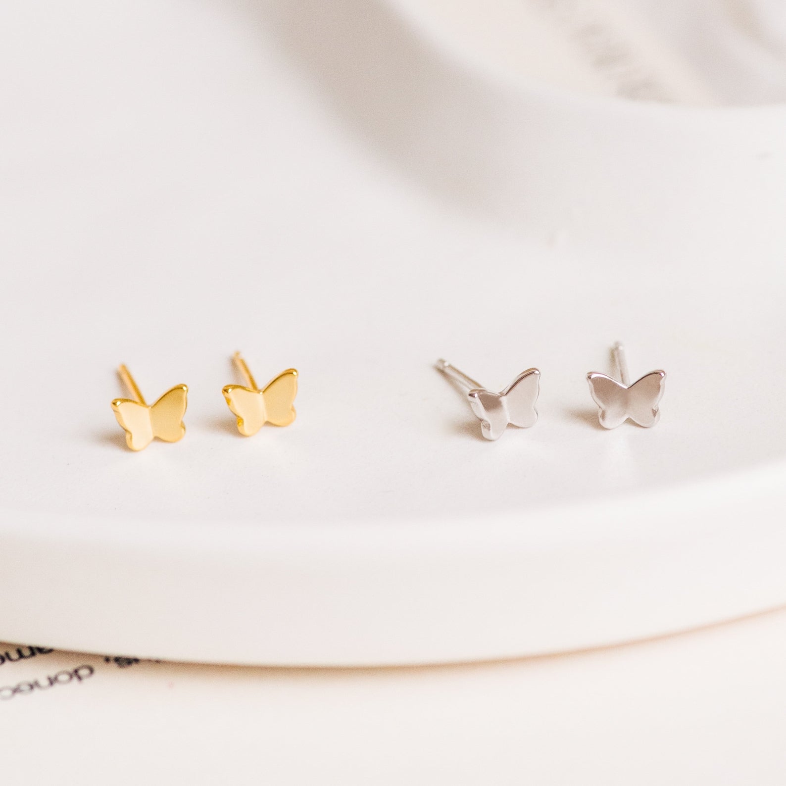 The Emilia Butterfly Studs feature two pairs of dainty butterfly earrings—one gold, one silver—displayed on a white surface, perfect for adding a trendy touch to any outfit.