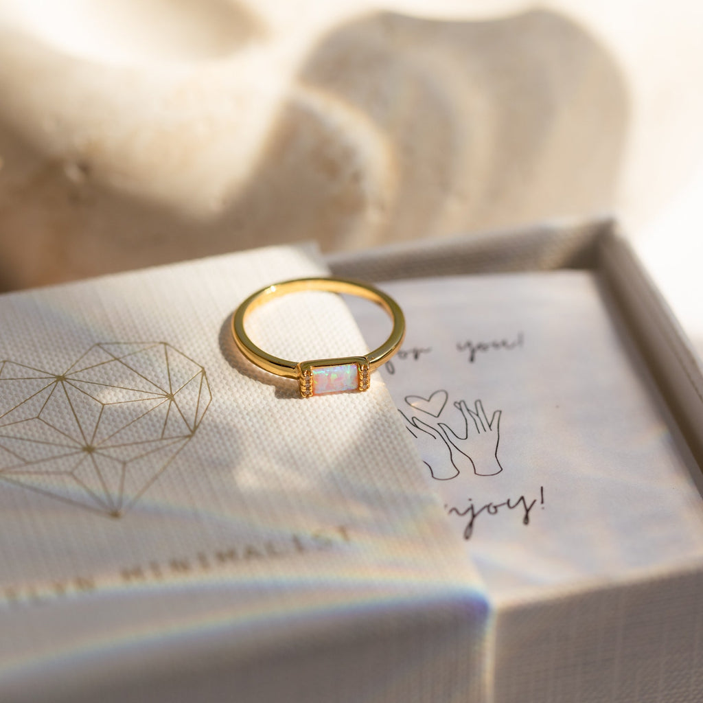 The Kiera Opal Baguette Ring, featuring a delicate opal stone, rests on a textured jewelry box with a geometric heart design—perfect as a unique promise ring.