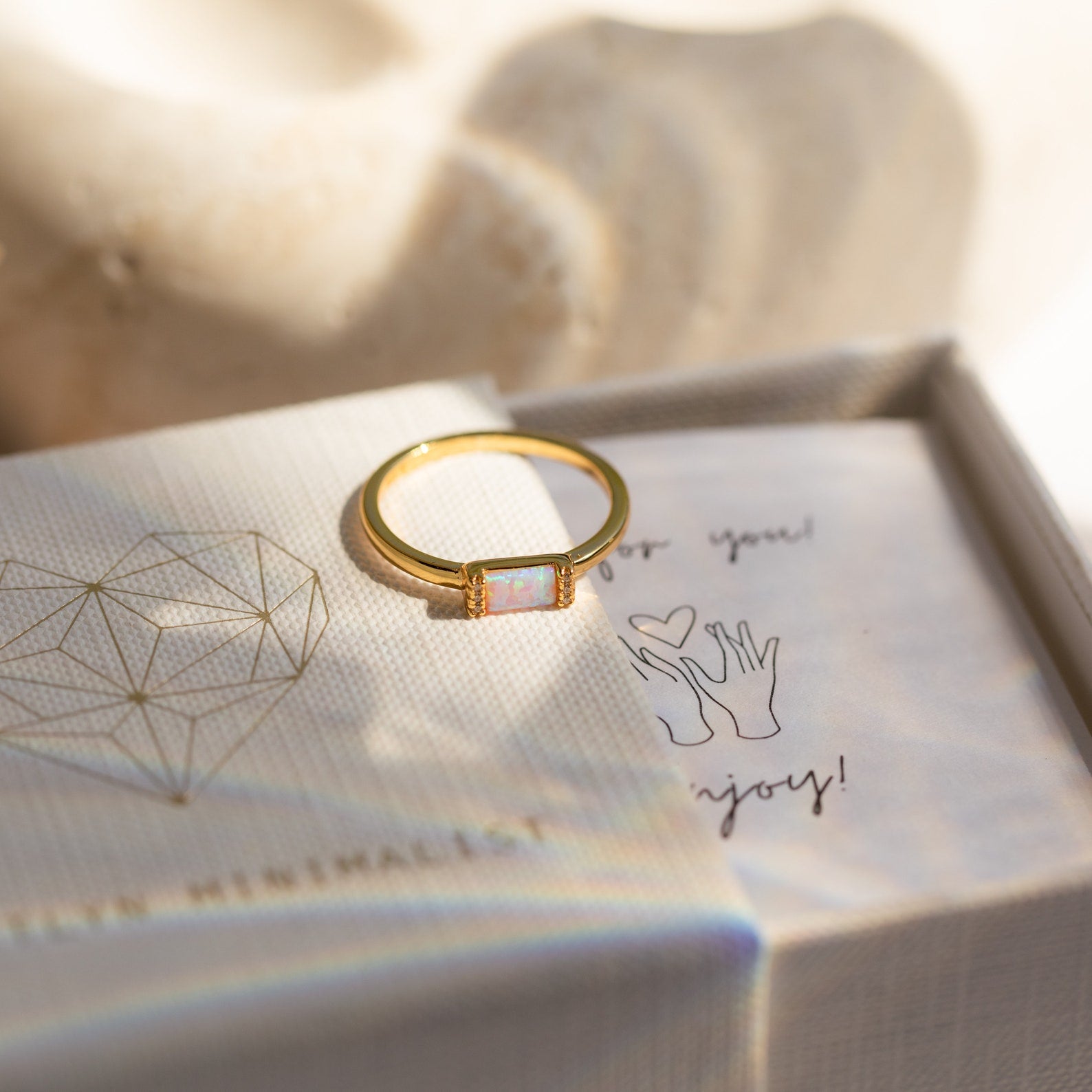 The Kiera Opal Baguette Ring, featuring a delicate opal stone, rests on a textured jewelry box with a geometric heart design—perfect as a unique promise ring.