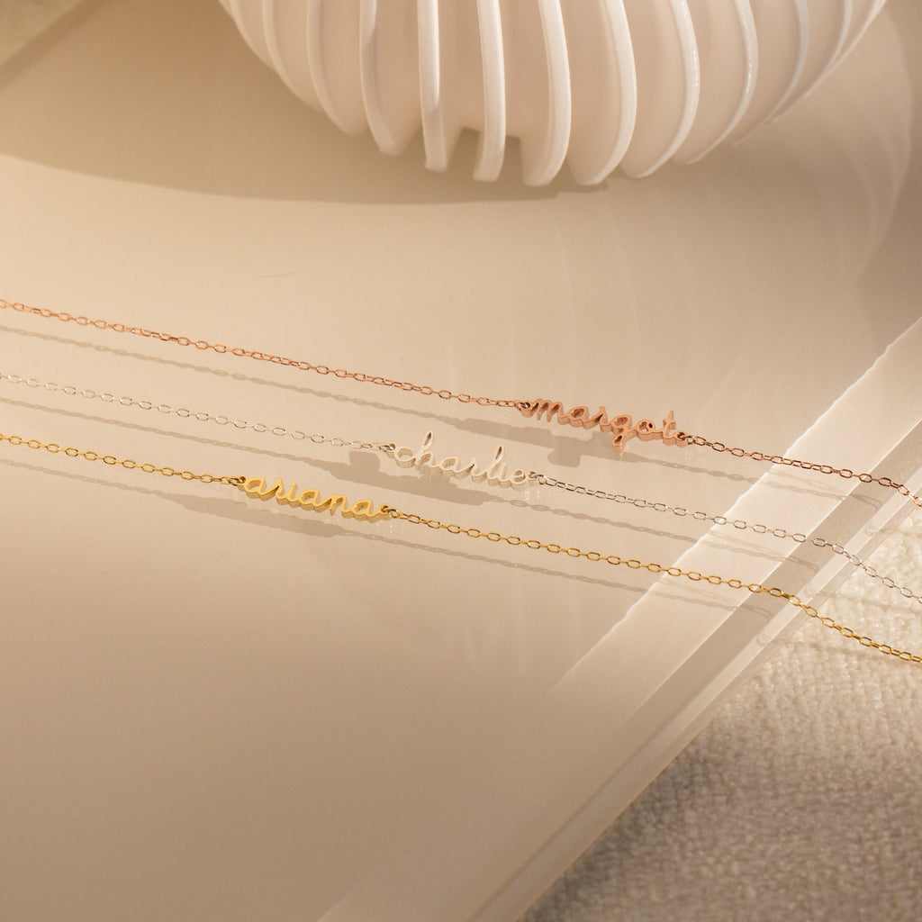 Three delicate name necklaces—ariana, charlie, and megan—are displayed on a beige surface beside a white vase, perfectly paired with the Dainty Tiny Name Bracelet for a chic minimalist look.