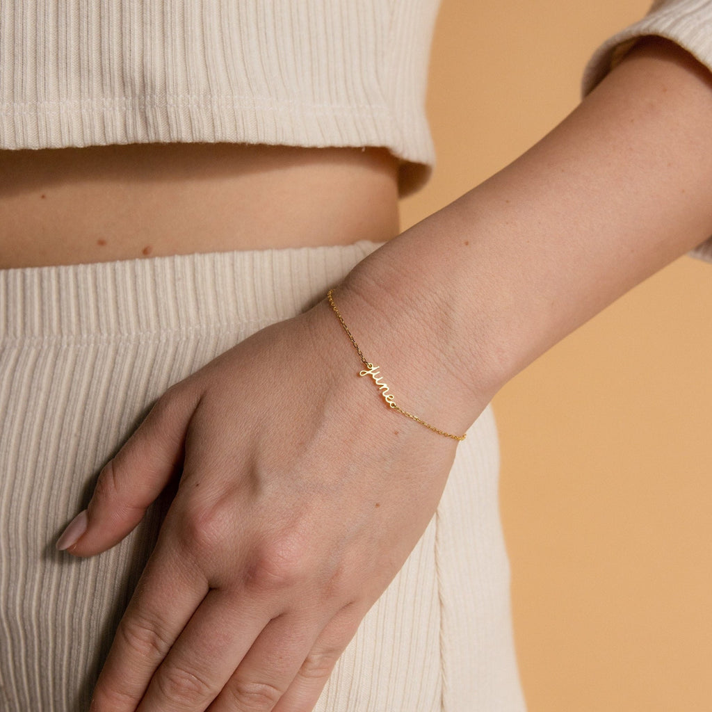 A hand rests on cream ribbed pants, showcasing the Dainty Tiny Name Bracelet—a minimal gold chain with a small personalized name pendant, adding an elegant and subtle touch.