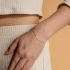 A hand rests on cream ribbed pants, showcasing the Dainty Tiny Name Bracelet—a minimal gold chain with a small personalized name pendant, adding an elegant and subtle touch.
