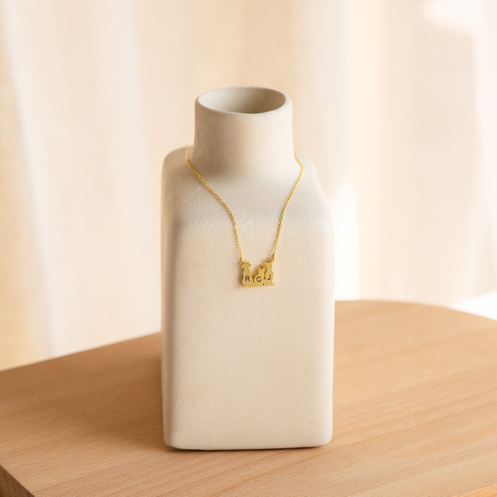 The Pet Initial Necklace, featuring gold letter charms, is beautifully displayed on a matte white ceramic vase—an ideal gift for pet lovers who cherish their furry friends.