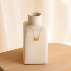 The Pet Initial Necklace, featuring gold letter charms, is beautifully displayed on a matte white ceramic vase—an ideal gift for pet lovers who cherish their furry friends.
