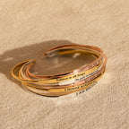 A stack of Custom Message Cuffs in gold and rose gold rests on a beige surface, each engraved bracelet perfect as a personalized gift for someone special.