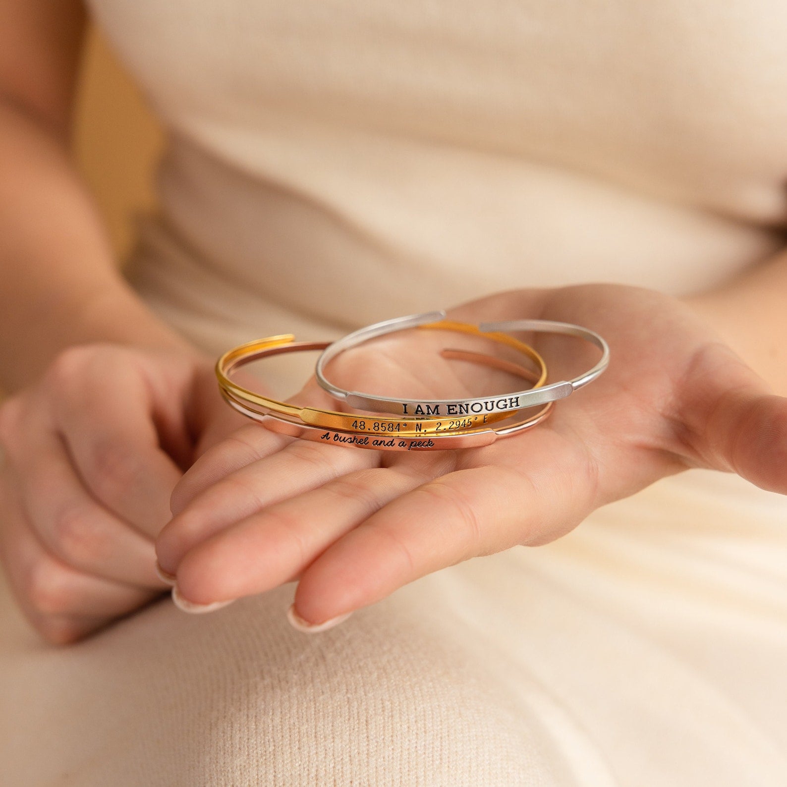 A hand holds three metal bangles, including the Custom Message Cuff engraved with "I AM ENOUGH" and numbers—a unique personalized gift.