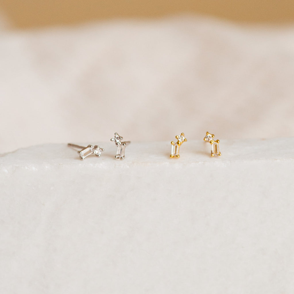 The Blair Baguette Diamond Drop Studs—two pairs in silver and gold—rest on a soft white surface, offering timeless style for minimalist earring lovers.