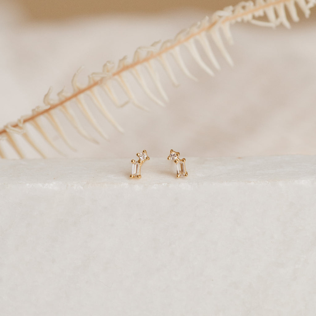 The Blair Baguette Diamond Drop Studs, elegant gold earrings with sparkling diamonds, are showcased on a white surface with a dried fern in the background.