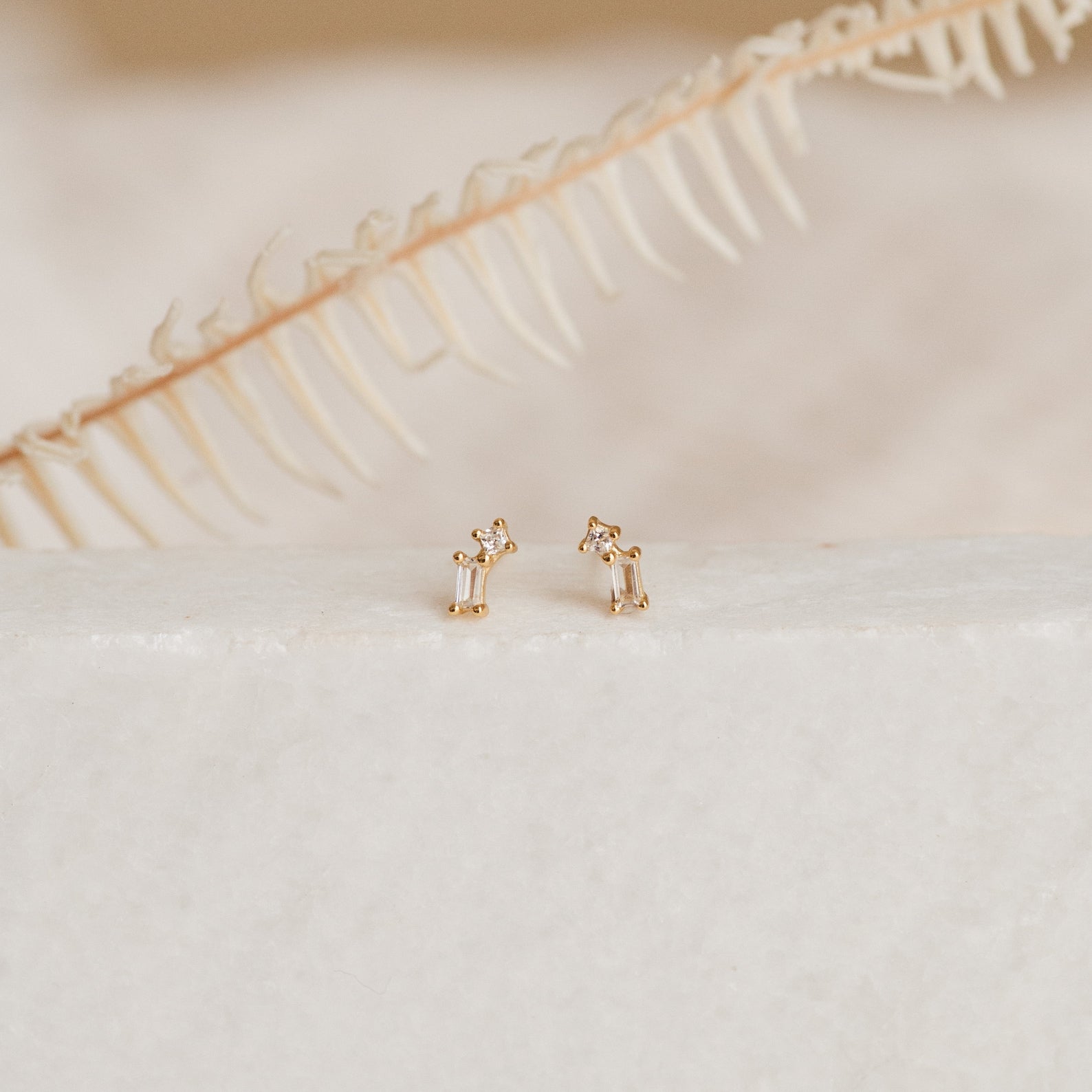 The Blair Baguette Diamond Drop Studs, elegant gold earrings with sparkling diamonds, are showcased on a white surface with a dried fern in the background.