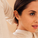 Close-up of a woman with blue eyes wearing a light beige turtleneck, showcasing our Hera Flower Stud earring on her earlobe, with her hair pulled back.