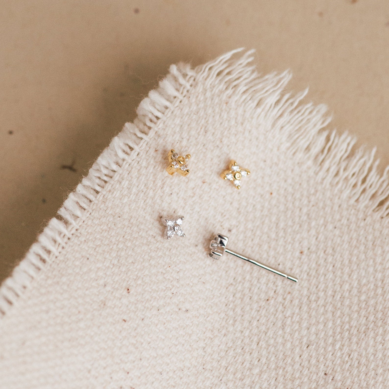 Four of our Hera Flower Stud earrings—two gold and two silver with clear stones—displayed on a textured cream fabric with frayed edges, laid on a neutral beige surface.