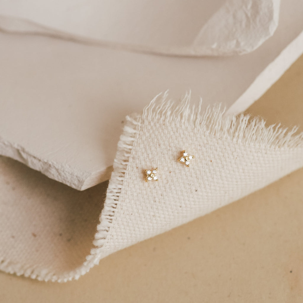 A pair of our Hera Flower Stud earrings in gold displayed on a piece of textured, frayed-edge cream fabric against a soft neutral background.