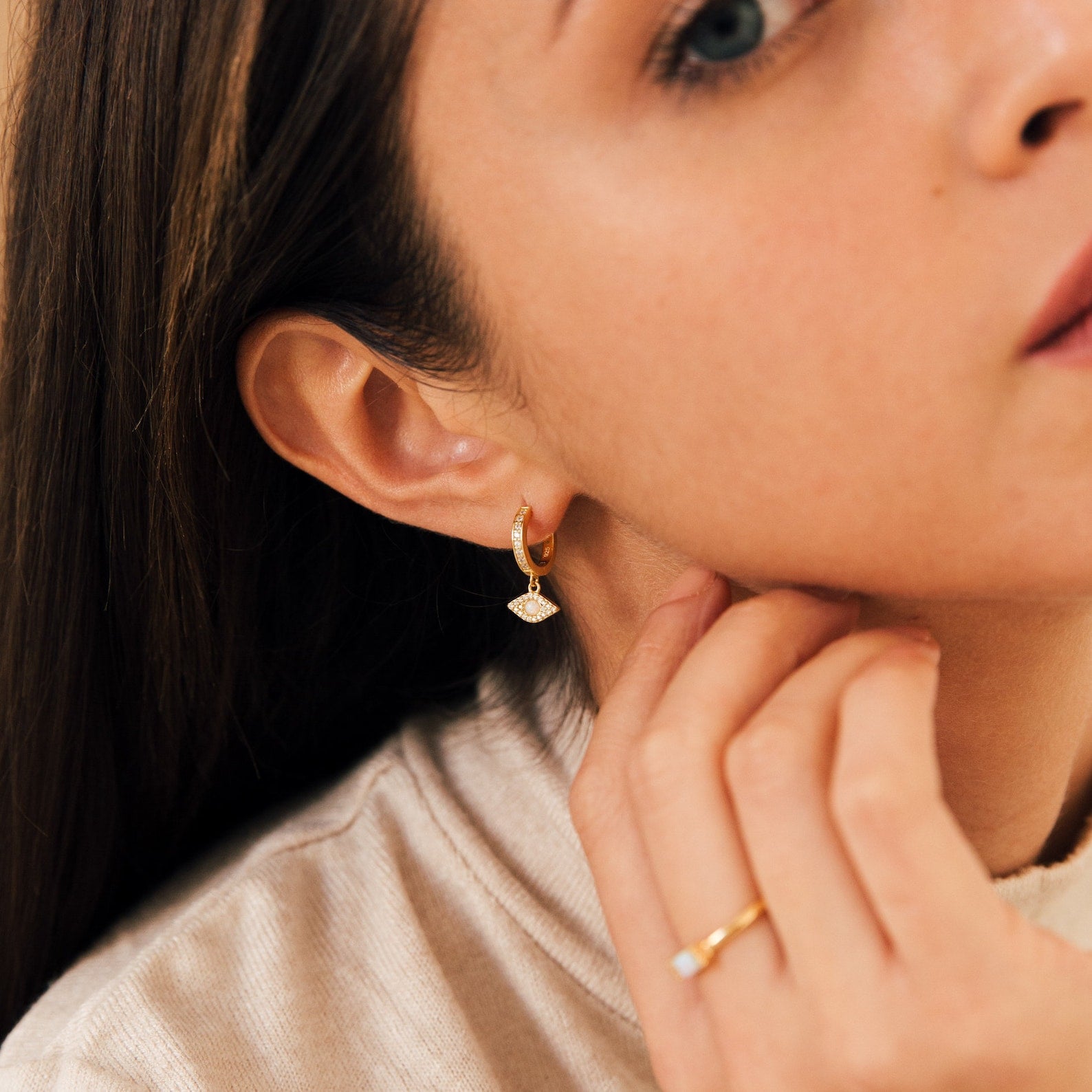 Woman wearing Aleah Opal Evil Eye Huggies and a gold ring, touching her neck and cheek.