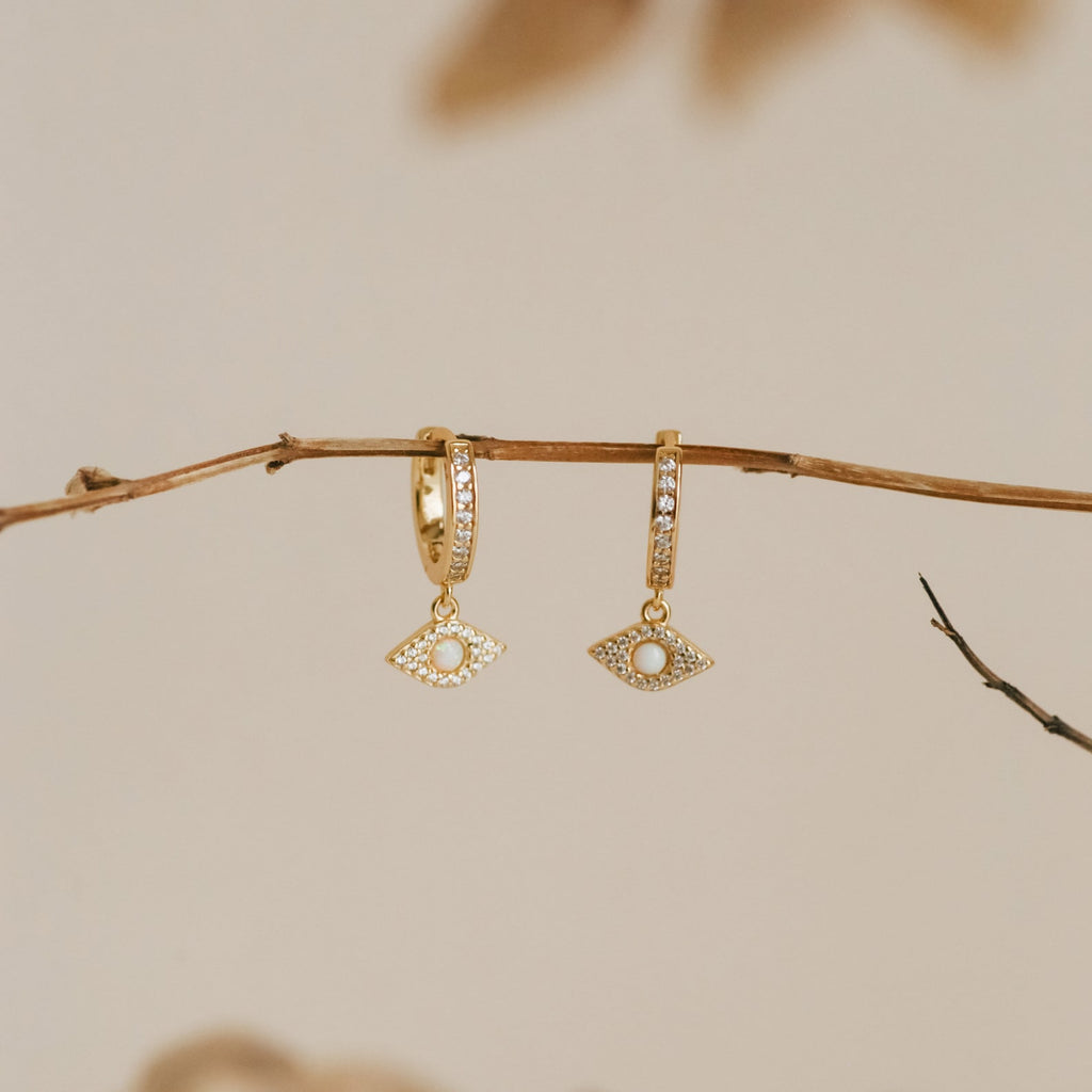 Aleah Opal Evil Eye Huggies—gold huggie hoop earrings featuring dangling eye-shaped charms inspired by evil eye jewelry—displayed on a bare branch against a neutral background.
