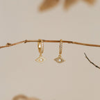 Aleah Opal Evil Eye Huggies—gold huggie hoop earrings featuring dangling eye-shaped charms inspired by evil eye jewelry—displayed on a bare branch against a neutral background.