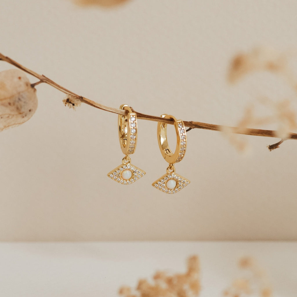 The Aleah Opal Evil Eye Huggies feature eye-shaped pearl and crystal charms, capturing the allure of evil eye jewelry and elegantly displayed against a beige background.