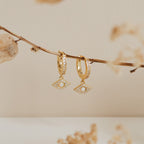 The Aleah Opal Evil Eye Huggies feature eye-shaped pearl and crystal charms, capturing the allure of evil eye jewelry and elegantly displayed against a beige background.