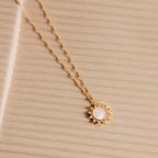 The Elia Sun Necklace features a gold sun-shaped pendant with a white mother of pearl center, displayed on textured beige—an eye-catching piece of boho-luxe jewelry.