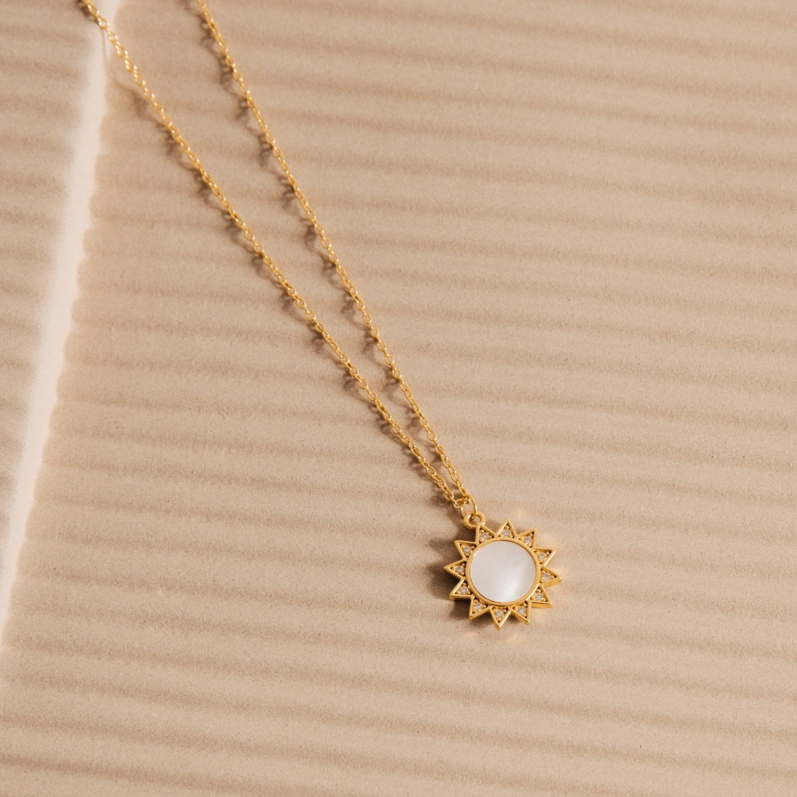 The Elia Sun Necklace features a gold sun-shaped pendant with a white mother of pearl center, displayed on textured beige—an eye-catching piece of boho-luxe jewelry.
