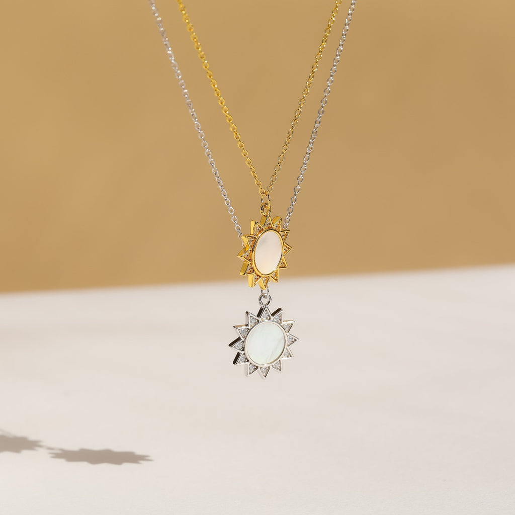 The Elia Sun Necklace features two sun-shaped pendants—one gold, one silver—on matching chains, styled against a beige background for a perfect boho-luxe jewelry statement.