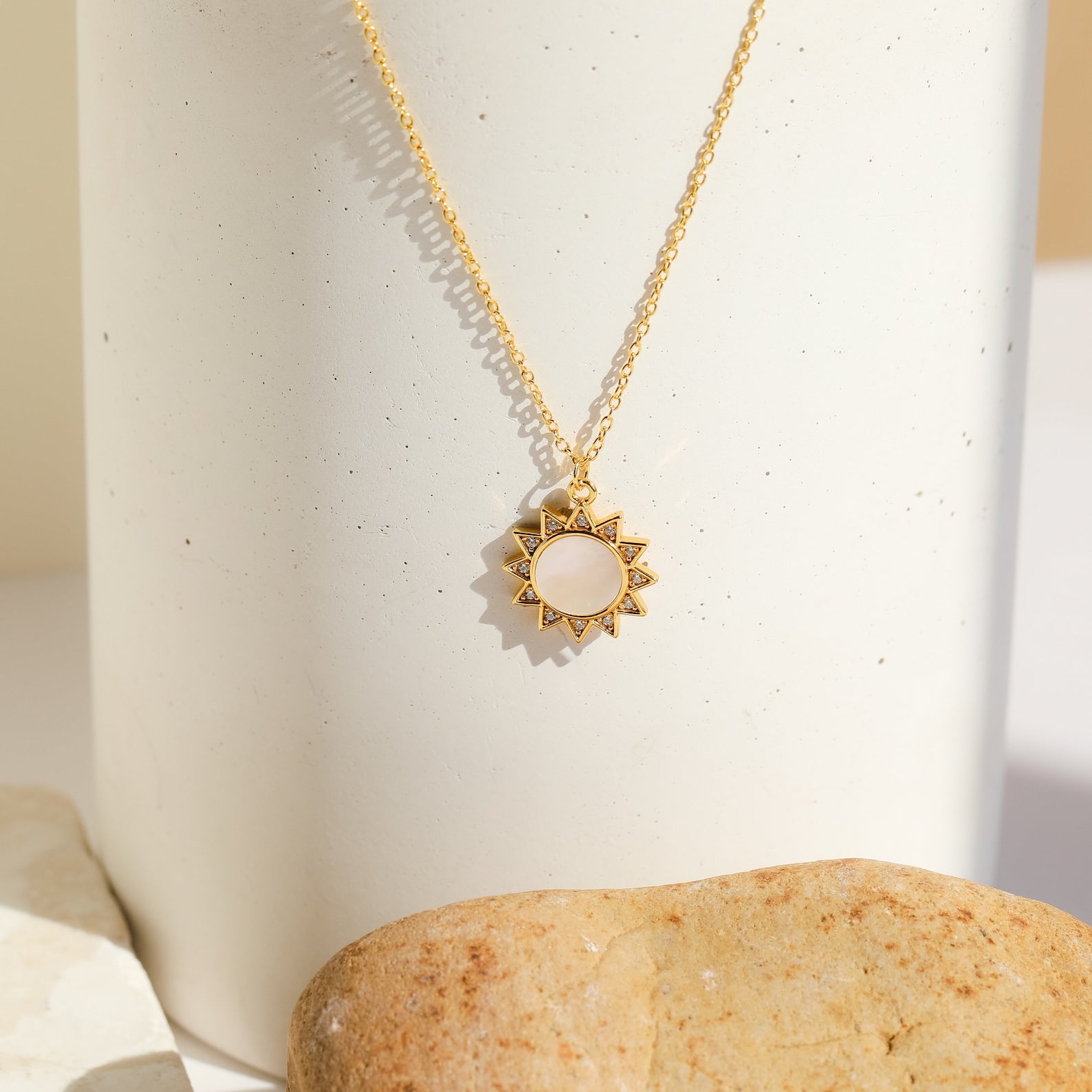 The Elia Sun Necklace features a gold sun-shaped pendant with a white mother of pearl center, shown on a light background with stones—radiating timeless boho-luxe charm.