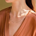 A woman wears the Dainty Pearl Station Necklace and a rust-colored V-neck blouse, sunlight illuminating her neck.