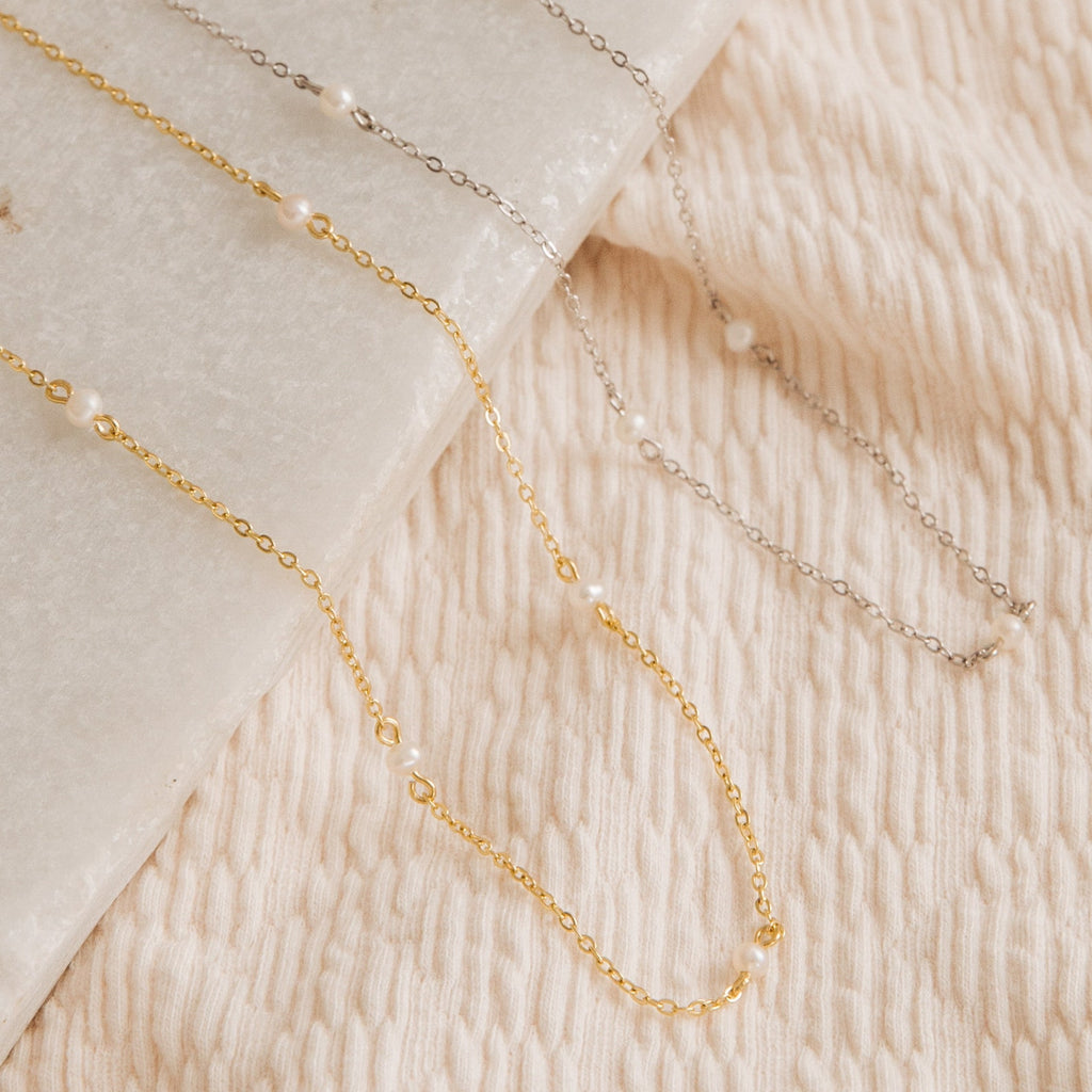 The Dainty Pearl Station Necklace, featuring gold and silver options with small pearls, is displayed on textured cream fabric and marble—offering a vintage style that makes an elegant gift for mom.
