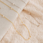 The Dainty Pearl Station Necklace, featuring gold and silver options with small pearls, is displayed on textured cream fabric and marble—offering a vintage style that makes an elegant gift for mom.