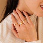 A woman's hand rests on her sweater, showcasing gold rings—including one with a pearl and the Jaida Peridot Duo Ring, featuring August's birthstone alongside other shimmering gemstone designs.