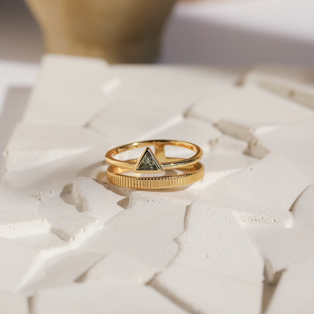 The Jaida Peridot Duo Ring, featuring two gold bands—one with a dark triangular stone and the other in Art Deco style—is displayed on a textured white surface.