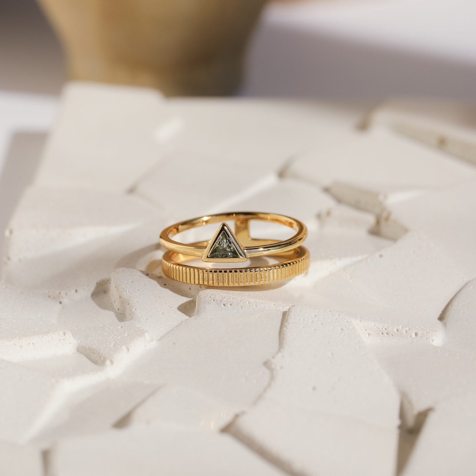 The Jaida Peridot Duo Ring, featuring two gold bands—one with a dark triangular stone and the other in Art Deco style—is displayed on a textured white surface.