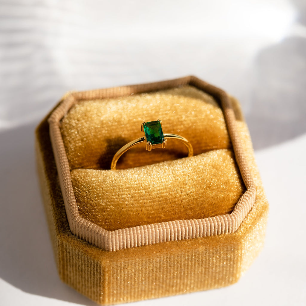 Harley Emerald Ring—an art deco gold ring with a rectangular green gemstone, presented in a yellow velvet hexagonal box and sunlit on a white surface. Ideal for vintage jewelry lovers.