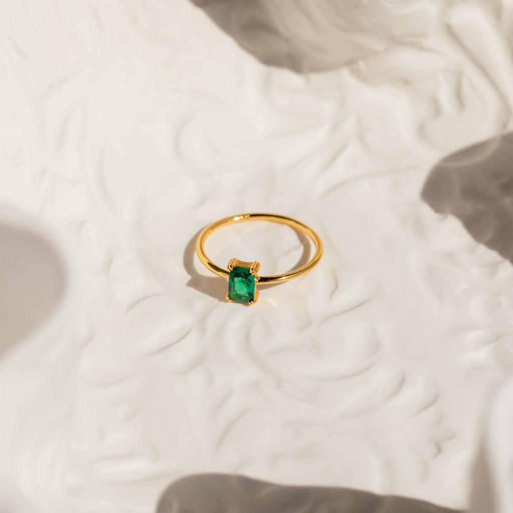 The Harley Emerald Ring features a rectangular green gemstone in a gold art deco setting, displayed on a textured white surface with soft shadows—a striking piece of vintage-style jewelry.