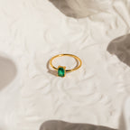The Harley Emerald Ring features a rectangular green gemstone in a gold art deco setting, displayed on a textured white surface with soft shadows—a striking piece of vintage-style jewelry.