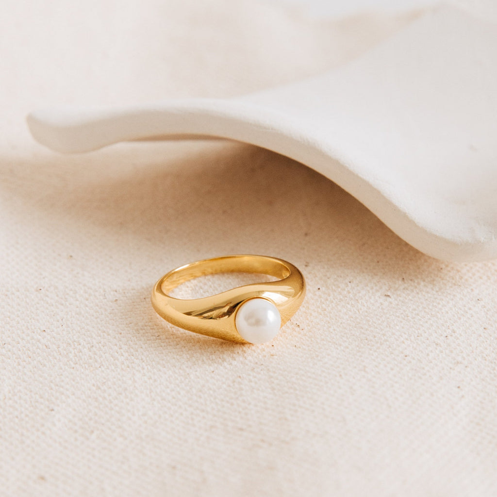 The Penelope Pearl Signet Ring, showcasing a single freshwater pearl, is displayed on textured beige fabric next to a white ceramic dish.
